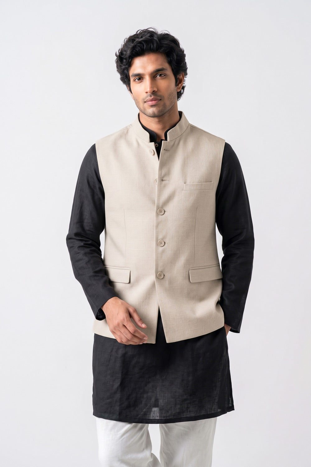 A man modeling a traditional Indian outfit consisting of a beige linen waistcoat (kurta) over a black long-sleeve shirt, paired with white trousers and white sneakers, against a plain light gray background.
