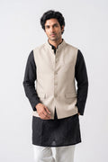 A male model wearing a beige linen Nehru-style vest with a stand collar, button closure, one chest pocket, and two flap pockets over a black kurta, paired with off-white trousers, against a light neutral background.