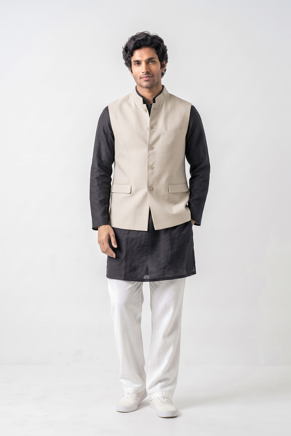 A man modeling a traditional Indian outfit consisting of a beige linen waistcoat (kurta) over a black long-sleeve shirt, paired with white trousers and white sneakers, against a plain light gray background.