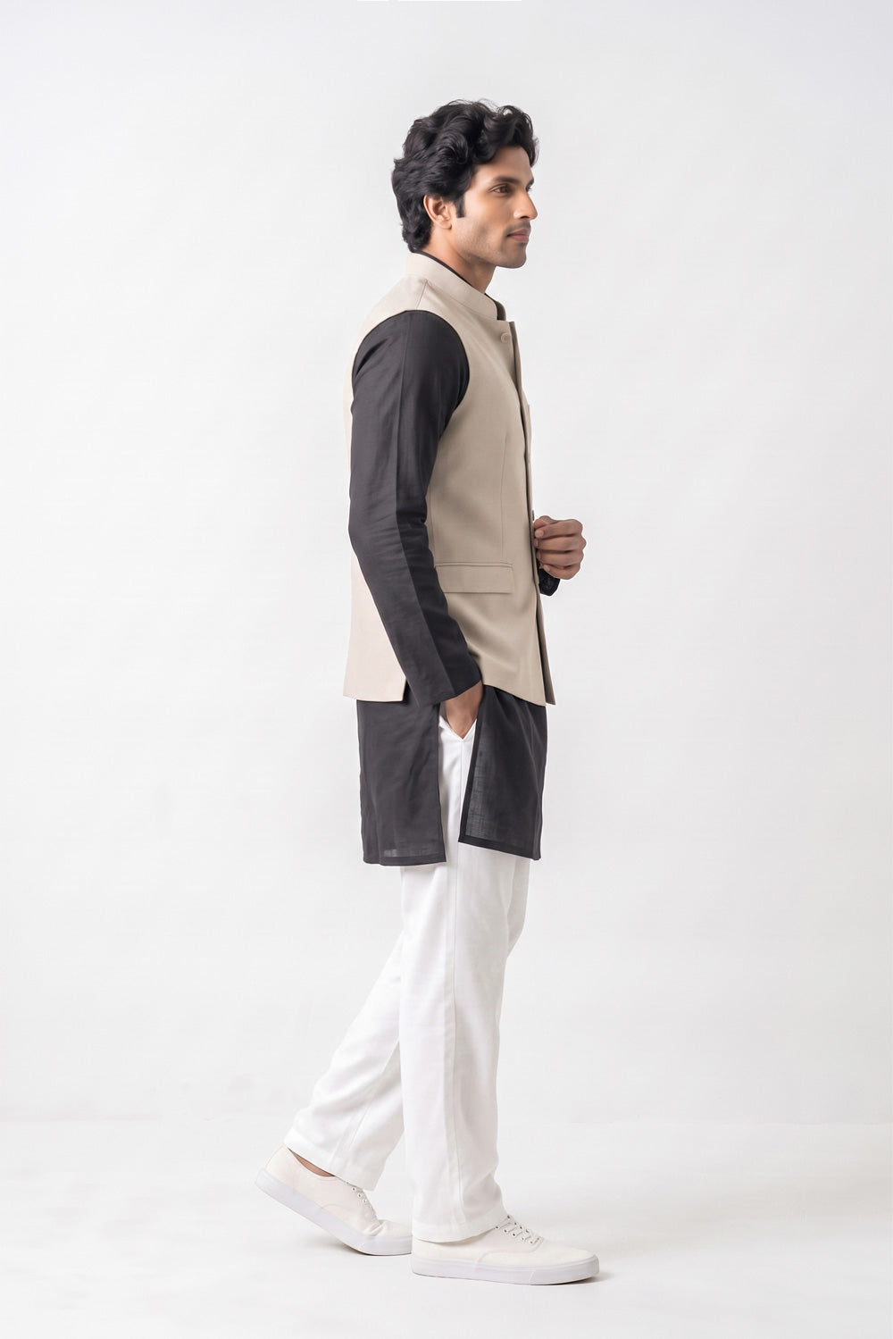 A man in profile wearing a beige sleeveless linen vest over a black long-sleeve kurta, paired with white trousers and white sneakers, standing against a plain white studio background.