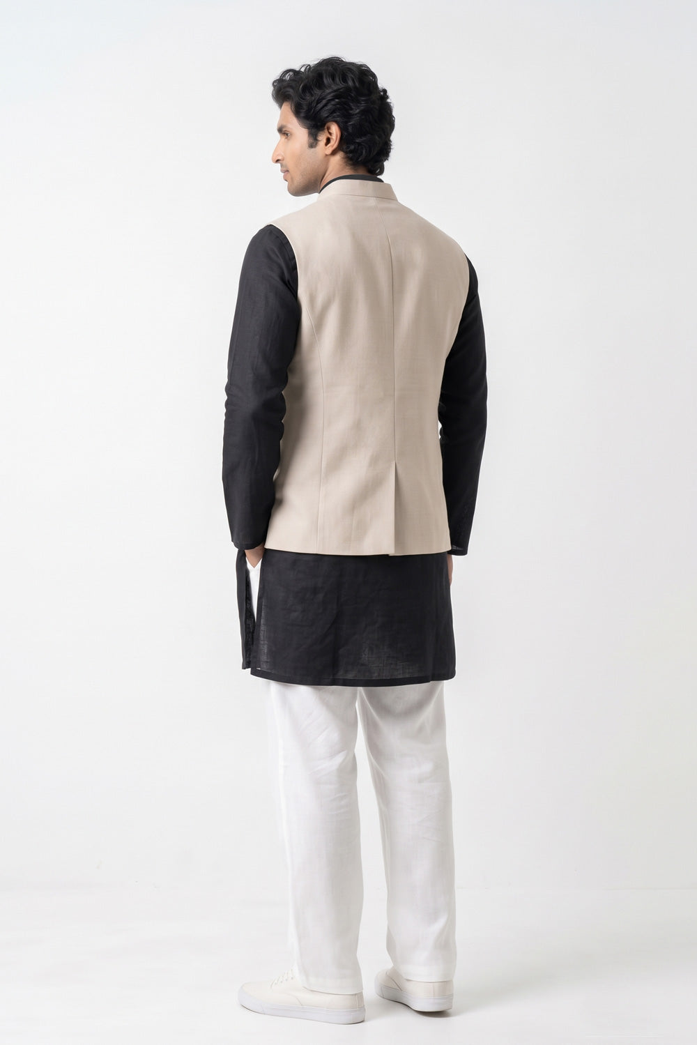 A man wearing a beige tailored linen waistcoat over a pale blue long-sleeve kurta, standing against a neutral studio background.