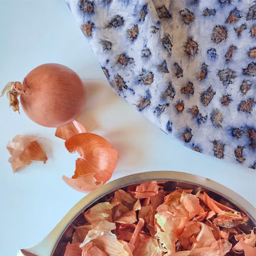 A stainless steel bowl filled with onion skins, with a whole onion and loose onion skins beside it on a white surface, and a blue-and-white patterned cloth in the upper right.