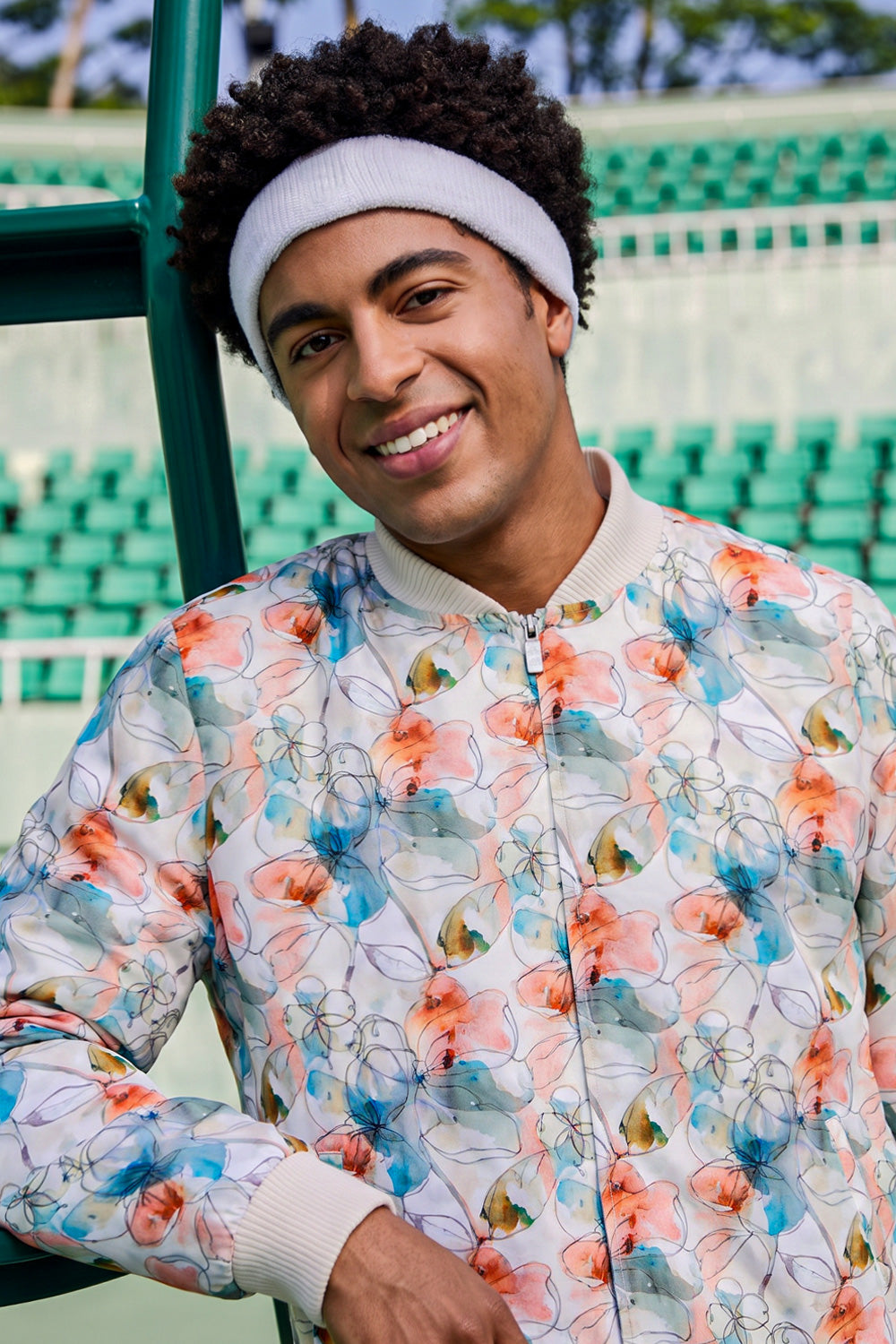 A smiling man wearing a white knit beanie and a white bomber hemp jacket with a colorful floral print, standing in front of green stadium seating.
