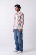 A man wearing a cream-colored bomber hemp jacket with a colorful floral print, blue jeans, and white sneakers, standing against a light gray studio background.