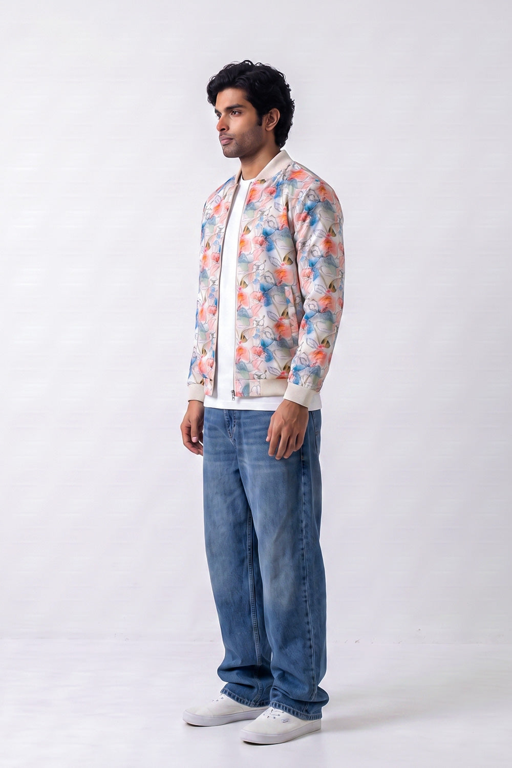 A man wearing a cream-colored bomber hemp jacket with a colorful floral print, blue jeans, and white sneakers, standing against a light gray studio background.