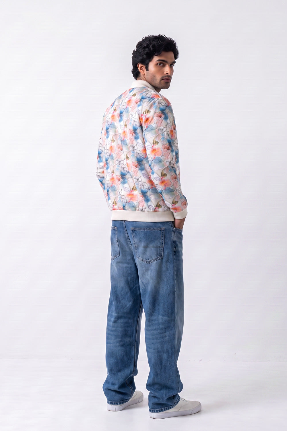 A man wearing a colorful floral print bomber Hemp jacket, blue jeans, and white sneakers, standing in profile against a plain white studio background.