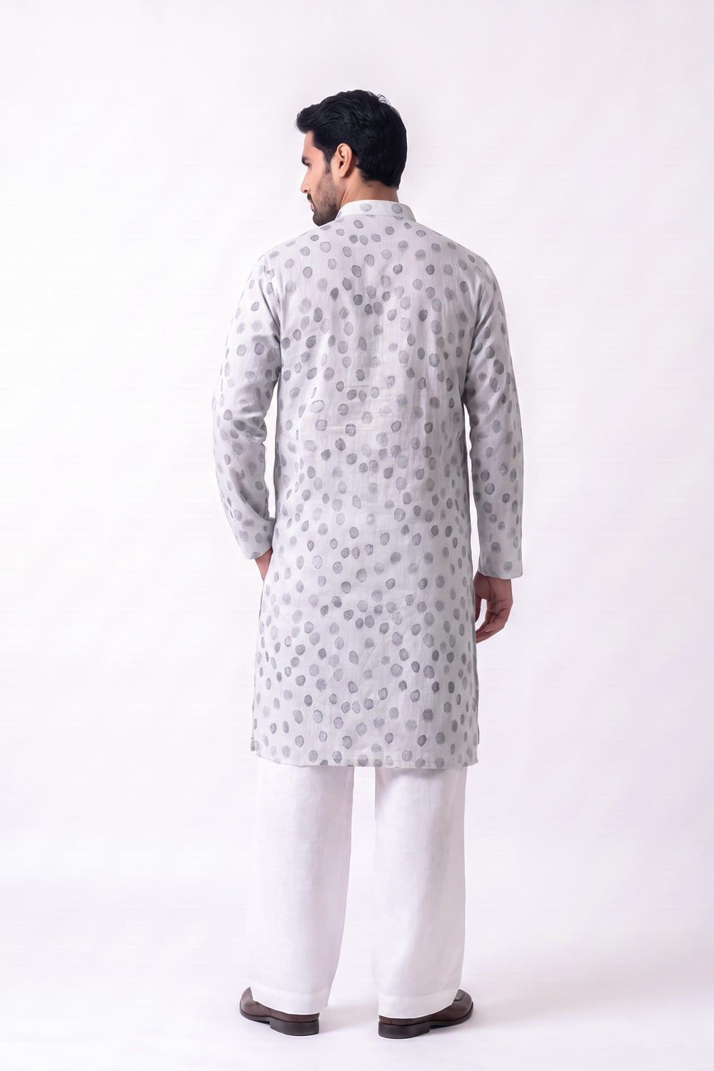 A man viewed from the back wearing a light gray long hemp kurta with a repeating circular pattern, paired with white trousers and dark brown shoes, against a plain white studio background.