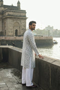 A man wearing a light gray and white patterned hemp kurta with white trousers stands on a stone walkway next to the water, with the Mumbai Gateway in the background.