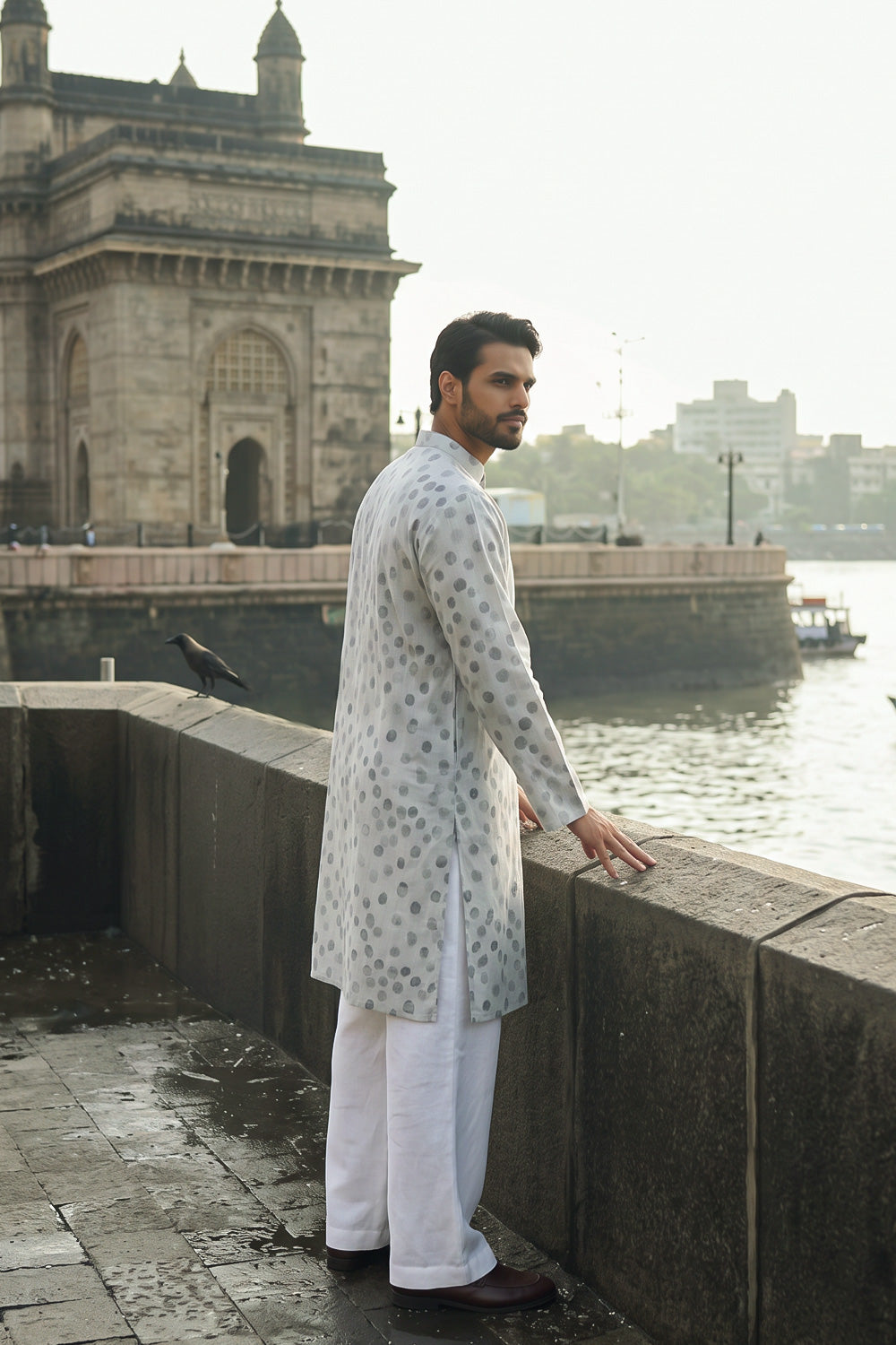 A man wearing a light gray and white patterned hemp kurta with white trousers stands on a stone walkway next to the water, with the Mumbai Gateway in the background.