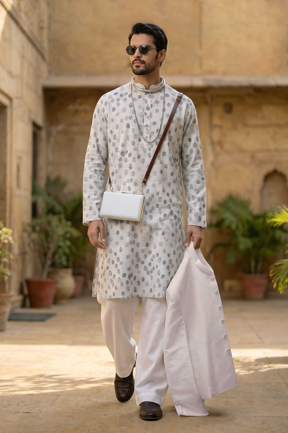 A man wearing a translucent white patterned hemp kurta with grey pebble-like motifs, paired with loose white trousers and brown loafers. He carries a white crossbody bag and holds a white blazer, accessorized with layered necklaces and round sunglasses. He stands in a warm, historic architectural courtyard with greenery.