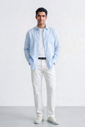 A man modeling a light blue linen button-down shirt over a white t-shirt, paired with white trousers and white sneakers, standing against a light gray studio background.