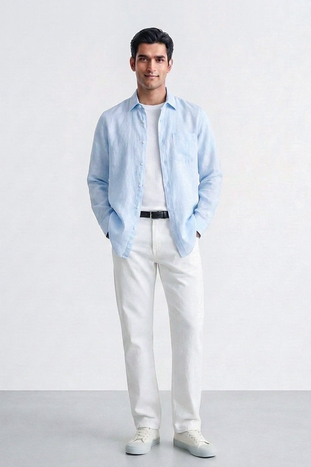 A man modeling a light blue linen button-down shirt over a white t-shirt, paired with white trousers and white sneakers, standing against a light gray studio background.