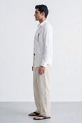 A man in profile wearing a white long-sleeve button-up linen shirt with a vertical chest pocket and beige linen trousers, standing against a plain light gray studio background.