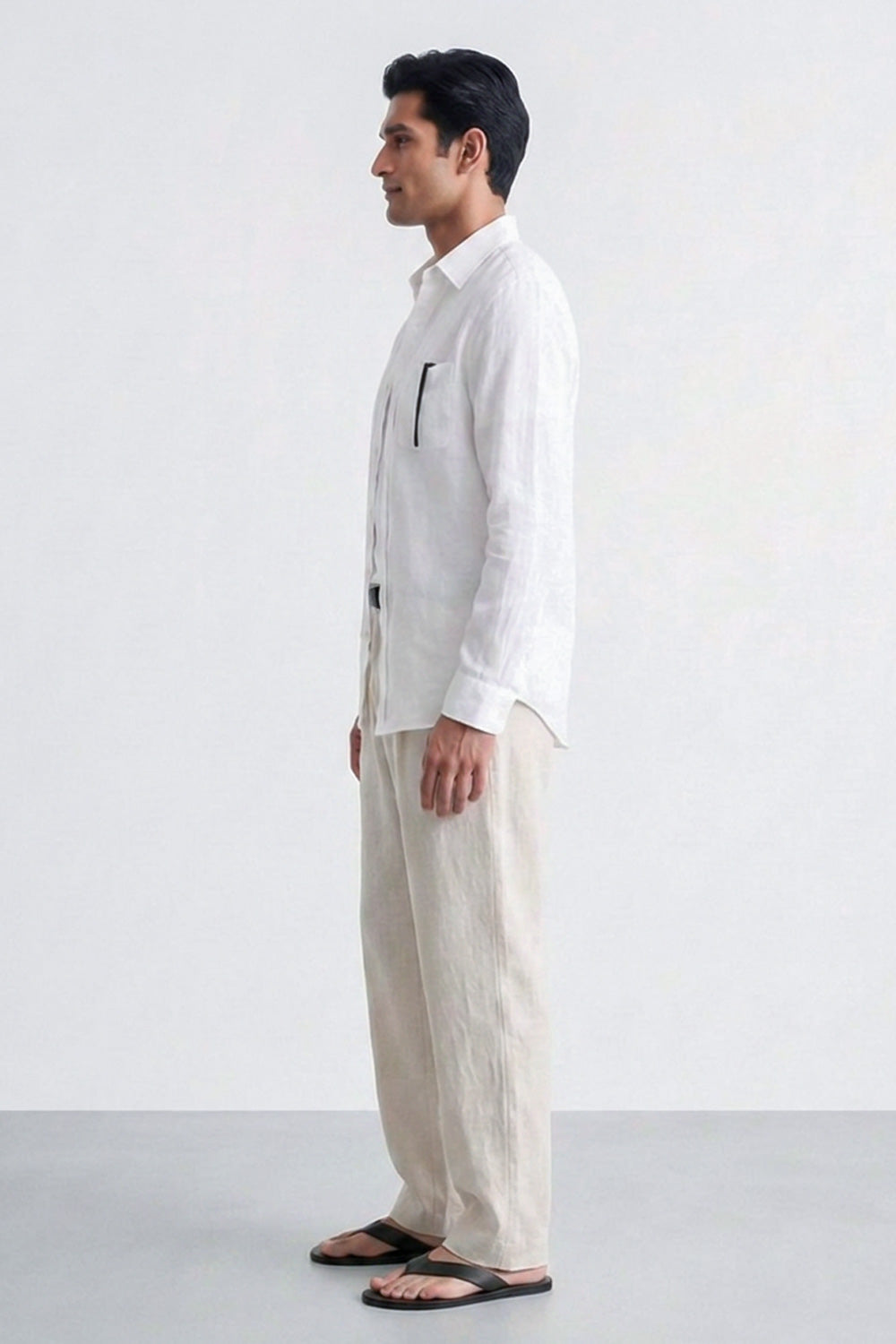 A man in profile wearing a white long-sleeve button-up linen shirt with a vertical chest pocket and beige linen trousers, standing against a plain light gray studio background.