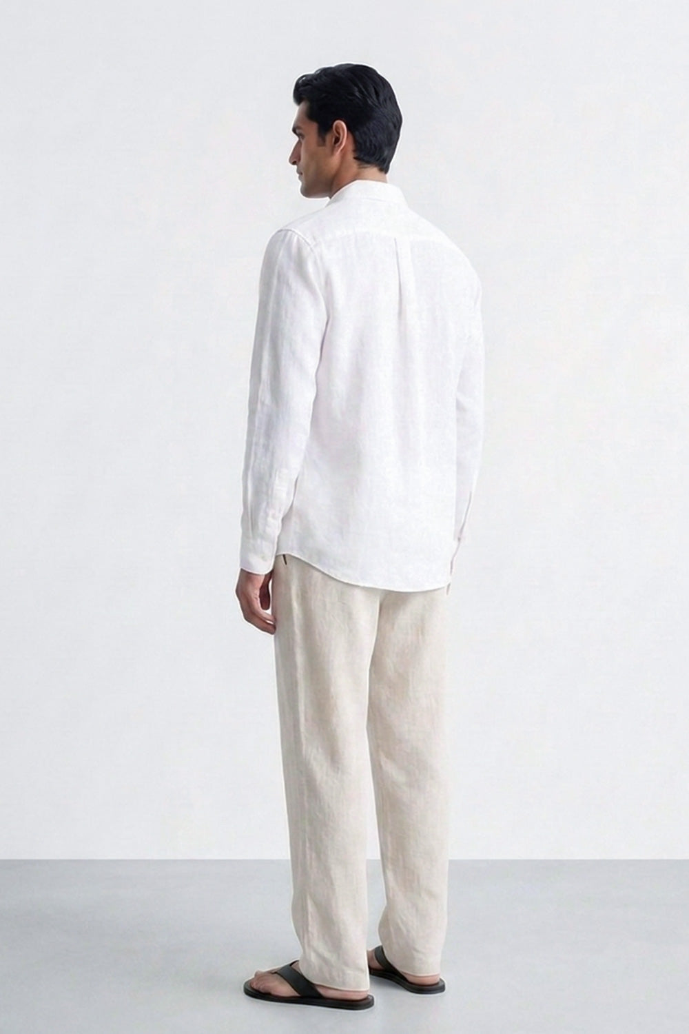 A man shown from the back wearing a white long-sleeve linen shirt and light beige linen trousers, standing against a plain white studio background.