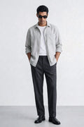 A man modeling a light gray linen button-down linen shirt over a white t-shirt, paired with dark gray trousers and black leather shoes, standing against a light gray studio background.