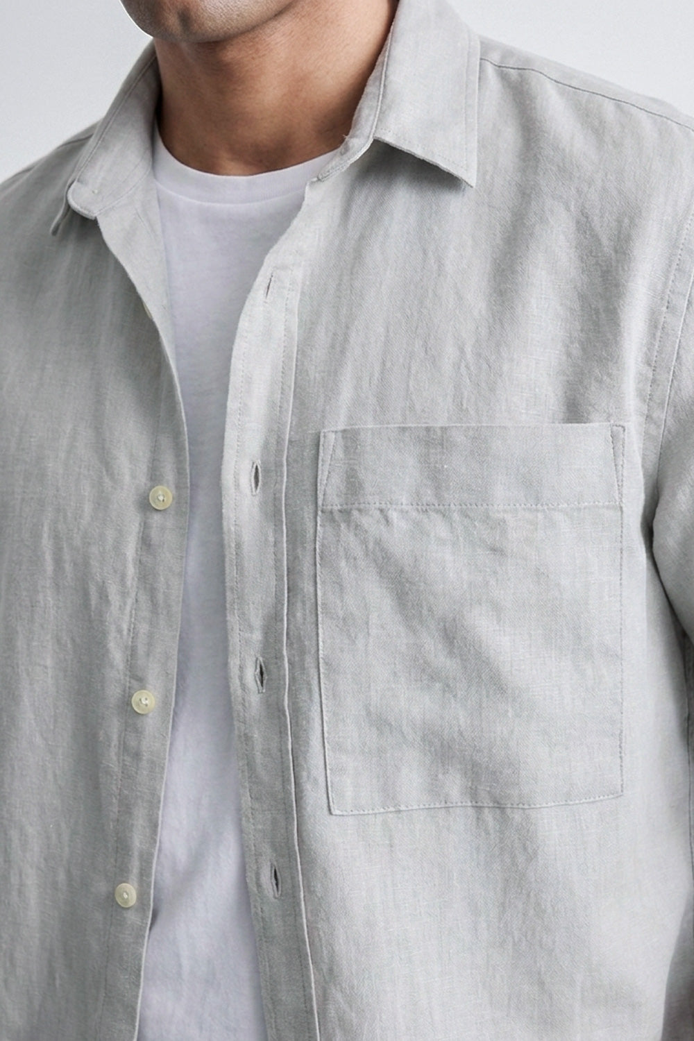 A close-up of a man wearing a light gray linen button-down shirt over a white t-shirt, showing the chest pocket and fabric texture.
