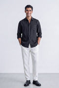 A man wearing a black long-sleeve linen button-down linen shirt and white trousers, standing against a light gray studio background.