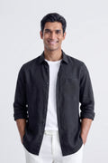 A man wearing a black long-sleeve linen button-up shirt over a white t-shirt and white pants, standing against a light gray studio background.