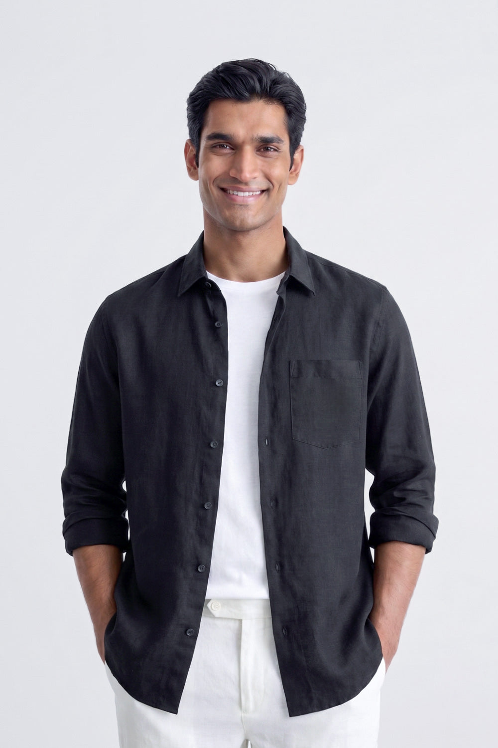 A man wearing a black long-sleeve linen button-up shirt over a white t-shirt and white pants, standing against a light gray studio background.