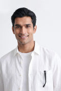 A smiling man with dark hair wearing a white button-down linen shirt over a white t-shirt, with a pair of black-framed glasses tucked into the chest pocket, against a plain white background.
