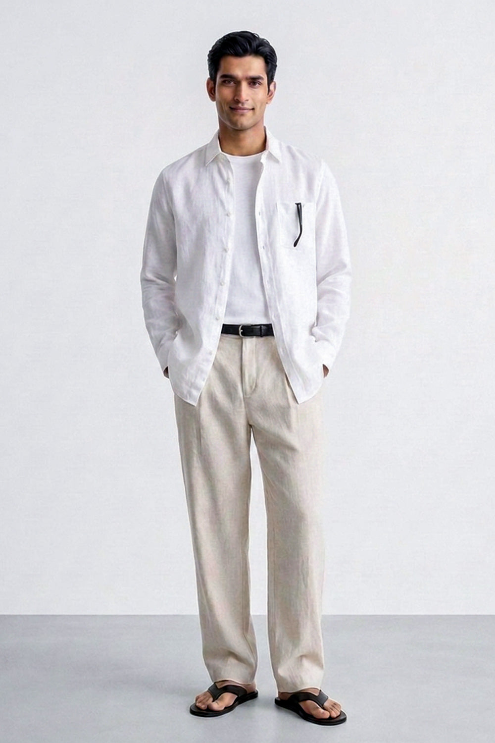 A man modeling a white long-sleeve linen shirt over a white tee, paired with beige linen trousers, a black belt, and black sandals, standing against a light gray studio background.