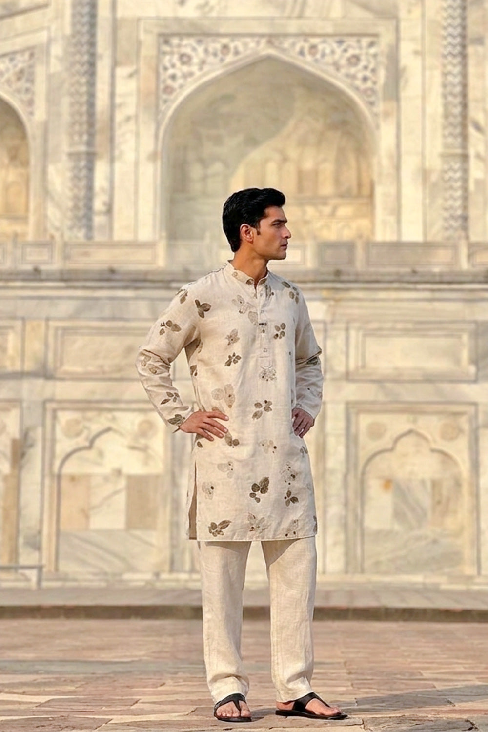 A man wearing a light beige kurta with brown floral patterns and matching trousers, standing in front of a white marble architectural background.