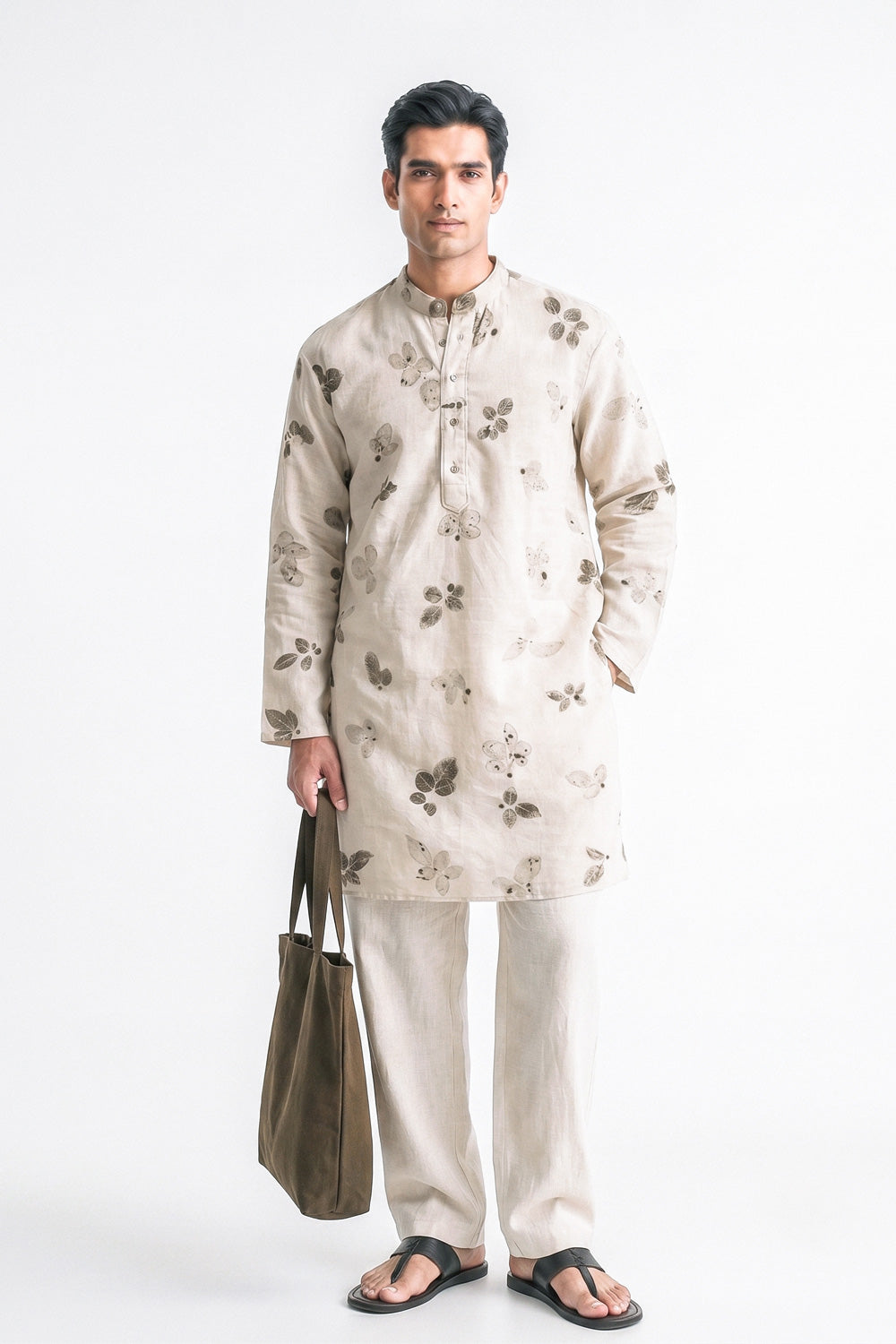 A man modeling a light beige kurta with a brown floral pattern, paired with matching beige trousers and black sandals, holding a brown tote bag against a plain white background.