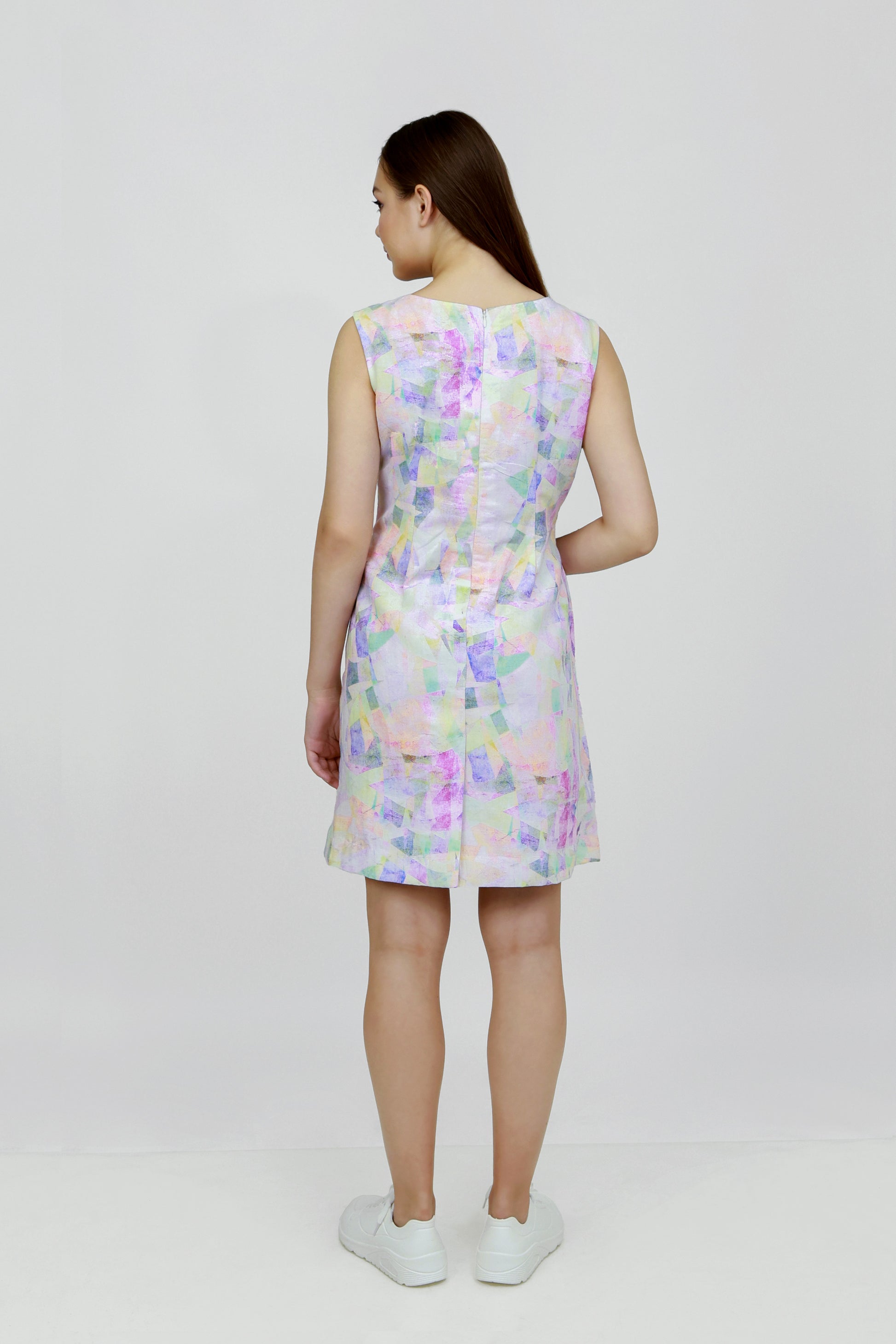 A woman standing in a studio setting, wearing a mid-length floral hemp seersucker dress with a boat neckline and sleeveless top, paired with white sneakers.