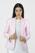 A woman standing outdoors wearing a pink linen blazer with a single-breasted design, peak lapel, and flap pockets. She also has on white trousers and is posing with her hands by her sides.