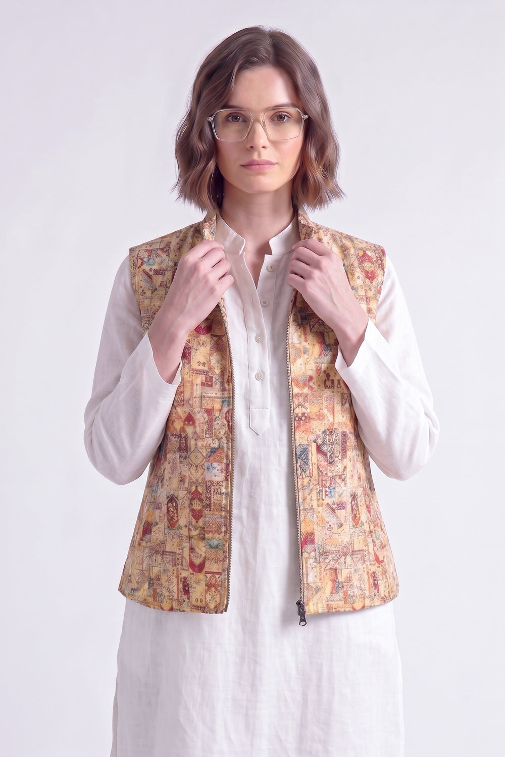 A woman wearing a white shirt and a beige  linen vest with a colorful, detailed traditional pattern, standing against a light gray background.