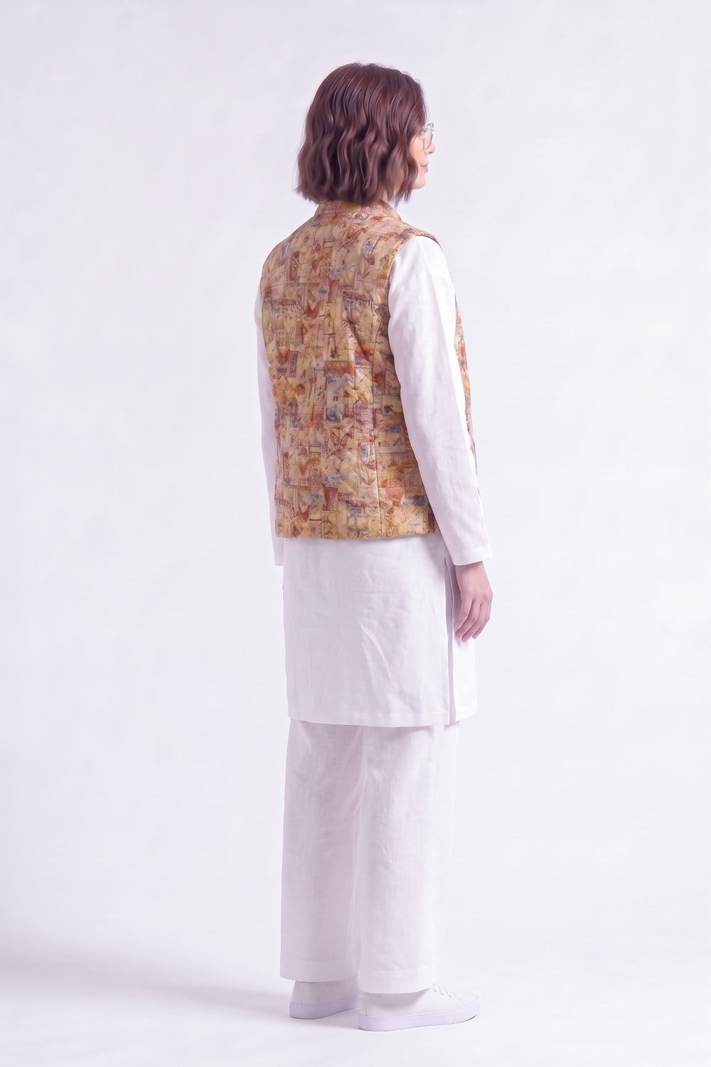 A model shown from the back wearing a white traditional outfit with a patterned mustard and brown linen vest, photographed against a plain white studio background.