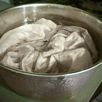 A close-up of a large hammered metal pot containing a light beige or off-white fabric bundle, likely for a traditional wash or steaming process.