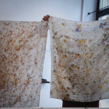 A person holds up two rectangular fabric panels with a mottled, speckled pattern in beige, brown, and muted gold tones against a plain indoor background.