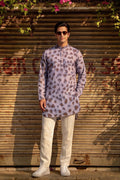 A man wearing a lavender and brown leopard-print kurta with cream trousers and white sneakers, standing in front of a closed wooden shutter door with hanging green leaves and pink flowers.