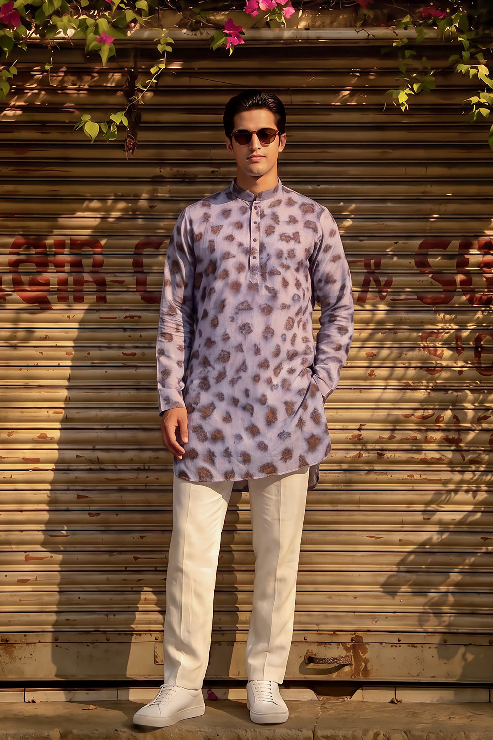 A man wearing a lavender and brown leopard-print kurta with cream trousers and white sneakers, standing in front of a closed wooden shutter door with hanging green leaves and pink flowers.