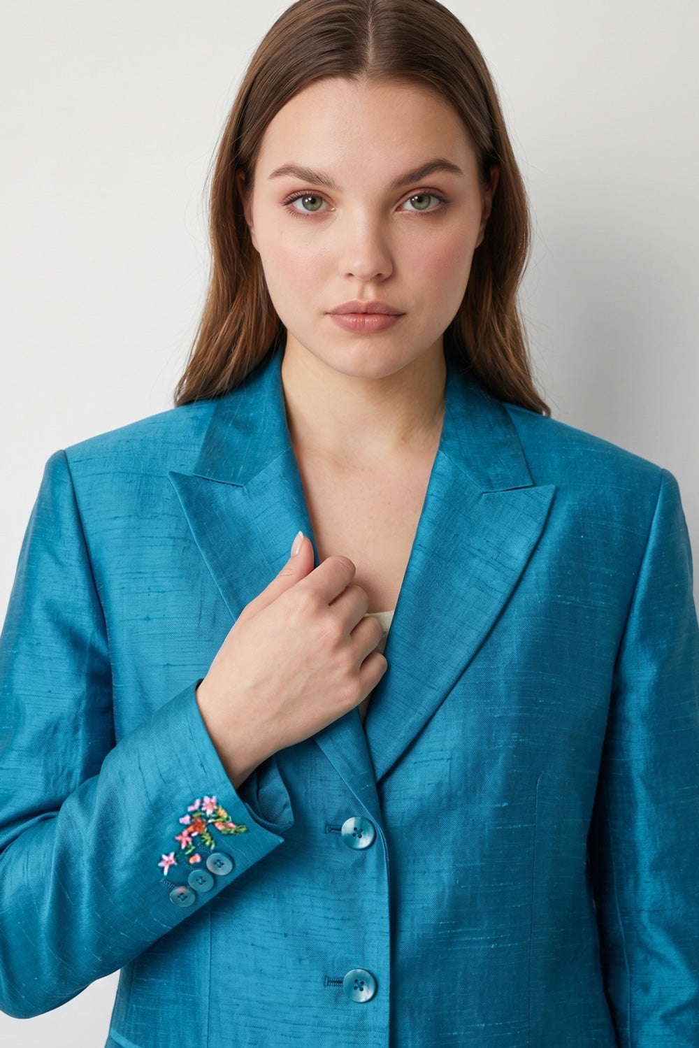 A woman stands against a light gray studio background wearing a bright turquoise-blue satin-like silk blazer and matching wide-leg trousers over a cream top and metallic sandals.
