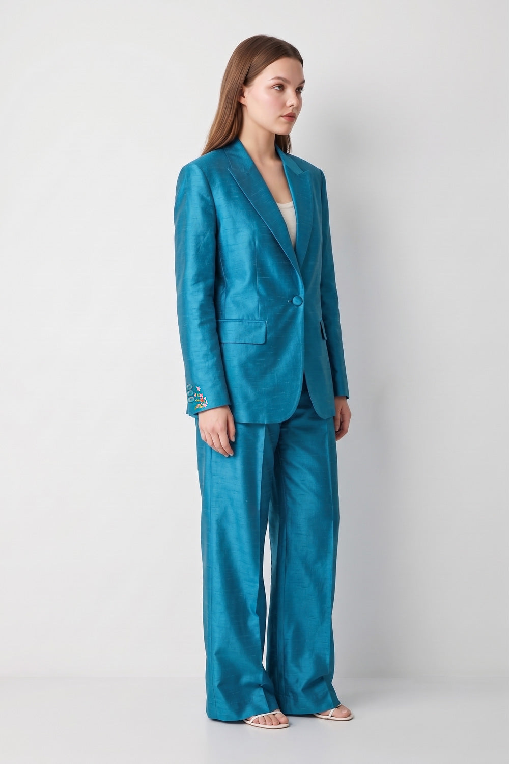 A woman modeling a matching teal blue satin-like silk blazer and wide-leg trousers set, standing against a light gray studio background.