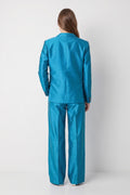 A model viewed from the back wearing a matching turquoise blue silk suit with a structured blazer and wide-leg trousers, set against a plain light gray studio background.