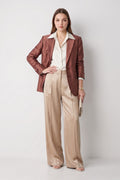 A model stands against a light gray studio background wearing a metallic bronze-brown silk blazer over a cream collared shirt, paired with wide-leg beige trousers and holding a small metallic clutch.