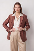 A woman modeling a rust-brown textured silk blazer over a cream collared shirt and beige trousers, standing against a light gray studio background.