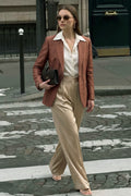 A woman walking on a city crosswalk wearing a rust-brown silk blazer, a cream button-up shirt, beige wide-leg trousers, and holding a black clutch bag.