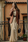 A woman stands on a balcony wearing a long cream satin dress with a brown silk blazer, a black belt, and holding a black handbag. The background features a warm terracotta-colored building wall and a black wrought-iron railing.