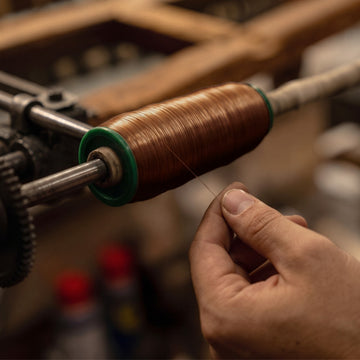 A close-up of a person's hand winding copper-colored thread onto a spool mounted on a machine in a workshop setting.