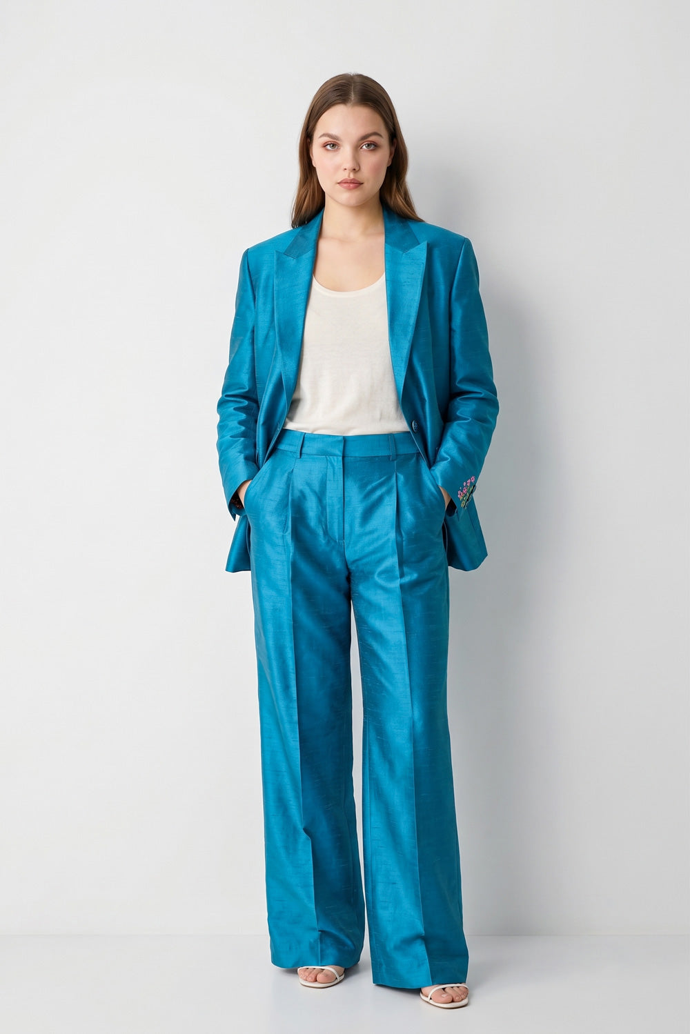 A woman stands against a light gray studio background wearing a bright turquoise-blue satin-like silk blazer and matching wide-leg trousers over a cream top and metallic sandals.