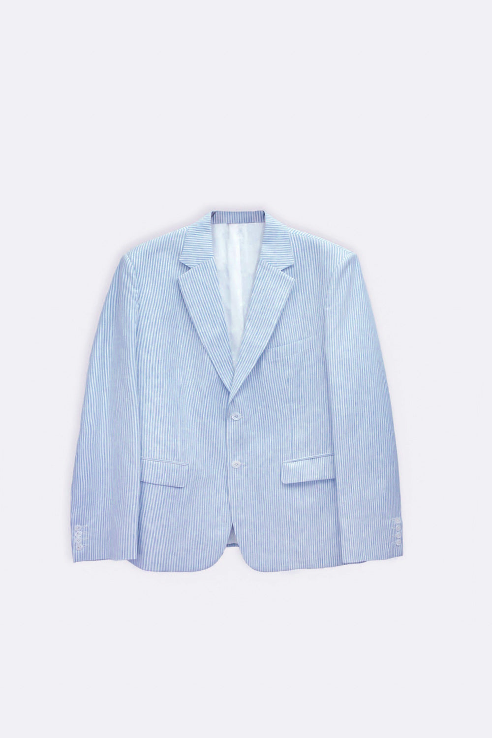 A man modeling a light blue linen blazer, white dress shirt, beige trousers, and white sneakers against a plain white studio background.
