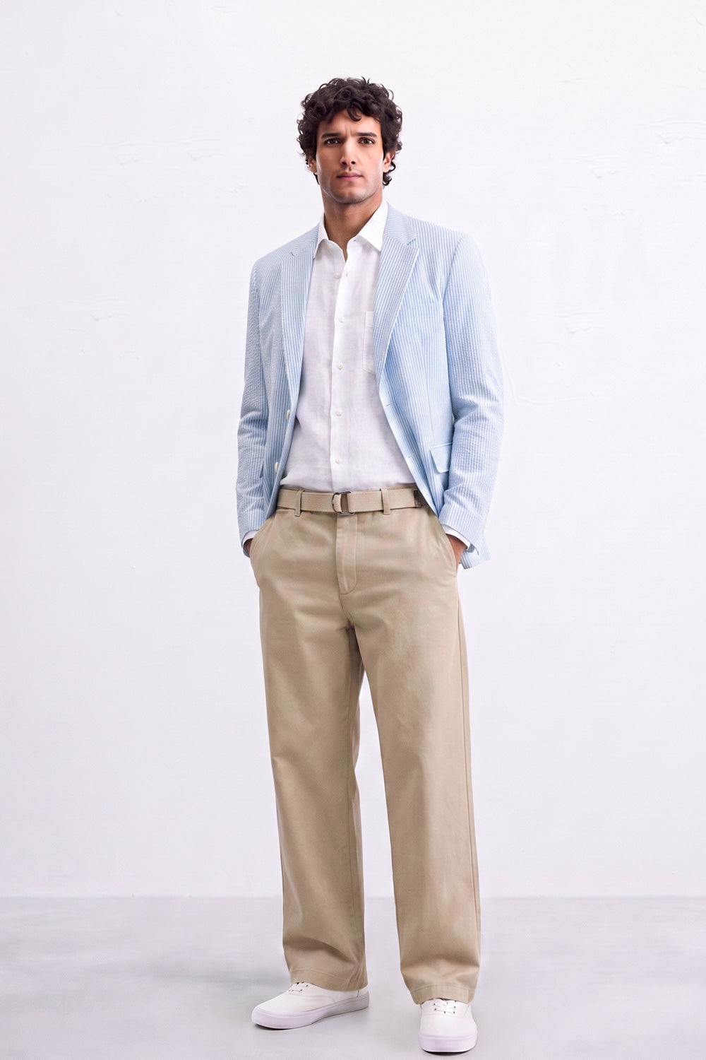 A man modeling a light blue linen blazer, white dress shirt, beige trousers, and white sneakers against a plain white studio background.