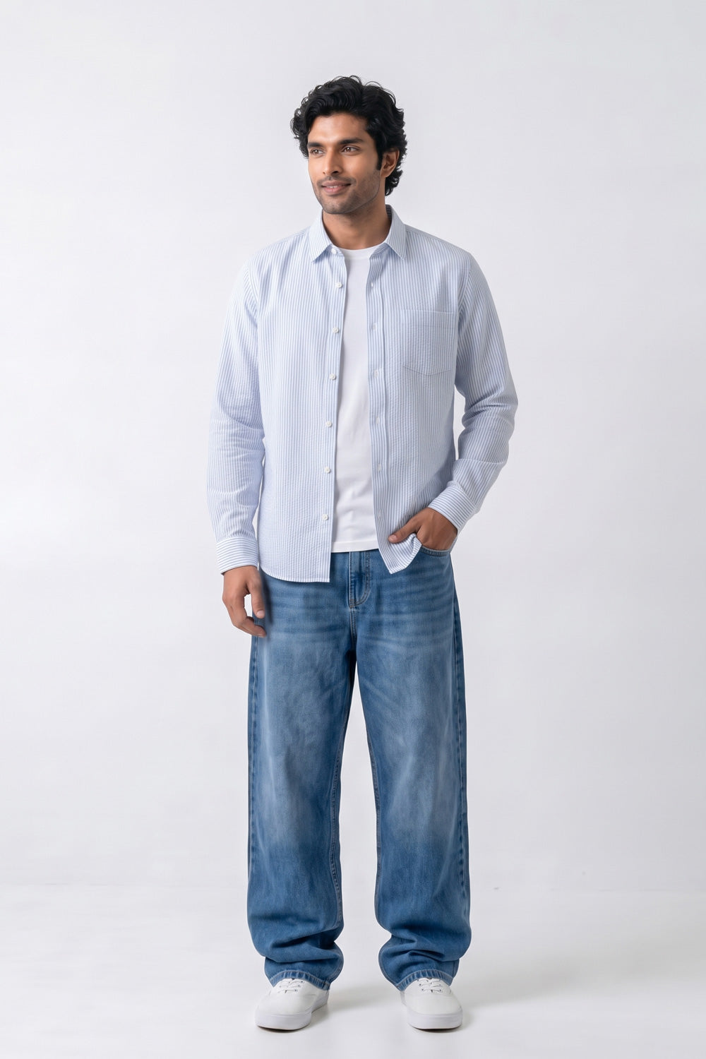 A man modeling a light blue long-sleeve button-up linen shirt over a white t-shirt, paired with blue jeans and white sneakers, against a plain white studio background.