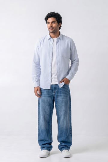 A man modeling a light blue long-sleeve button-up linen shirt over a white t-shirt, paired with blue jeans and white sneakers, against a plain white studio background.