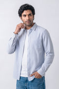 A man wearing a light blue and white vertically striped long-sleeve button-down linen shirt over a white t-shirt and blue jeans, standing against a light gray studio background.